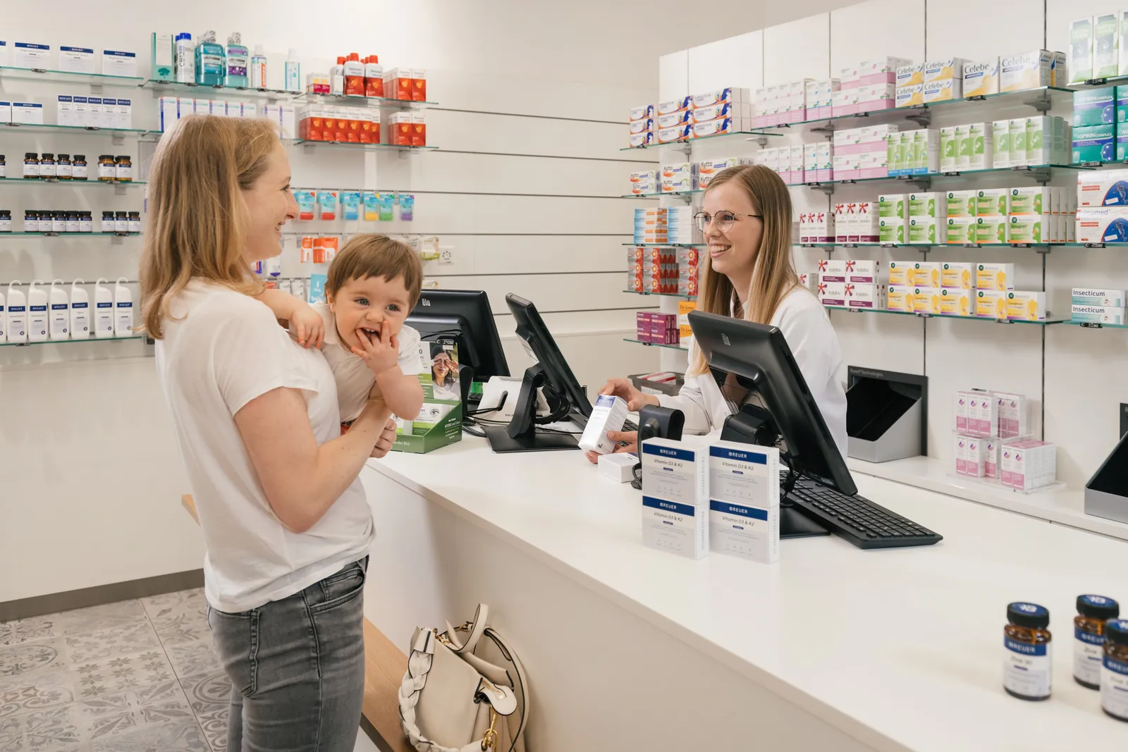Beratung in der Saggen-Apotheke: eine Mitarbeiterin lächelt an der Theke einer Mutter mit Kleinkind entgegen