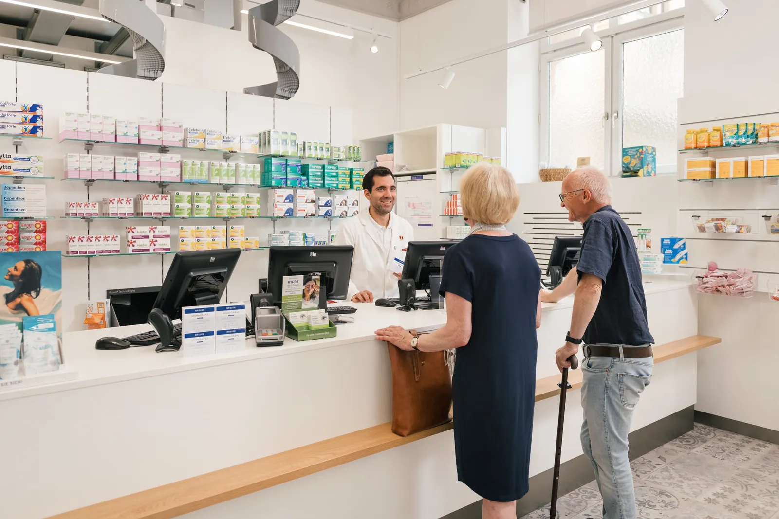 Ein Apotheker berät zwei Kundinnen und Kunden an der weißen Theke der Saggen-Apotheke