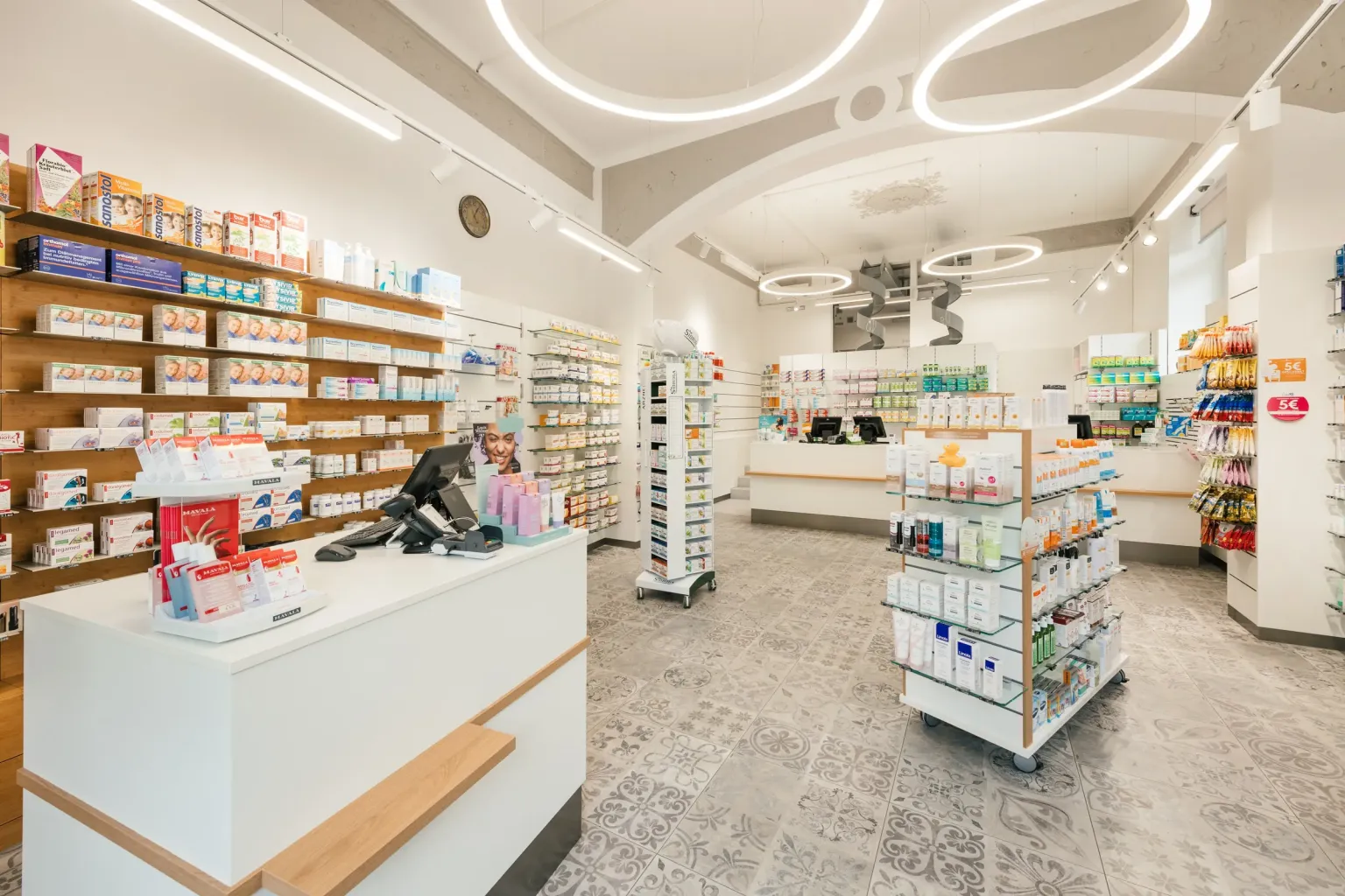 Heller Verkaufsraum der Saggen-Apotheke mit Gewölbedecke, Ringleuchten, Beratungstheken und Regalen